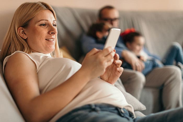 a female in brown having a online Telehealth consultation from home, while her partner is with kids
